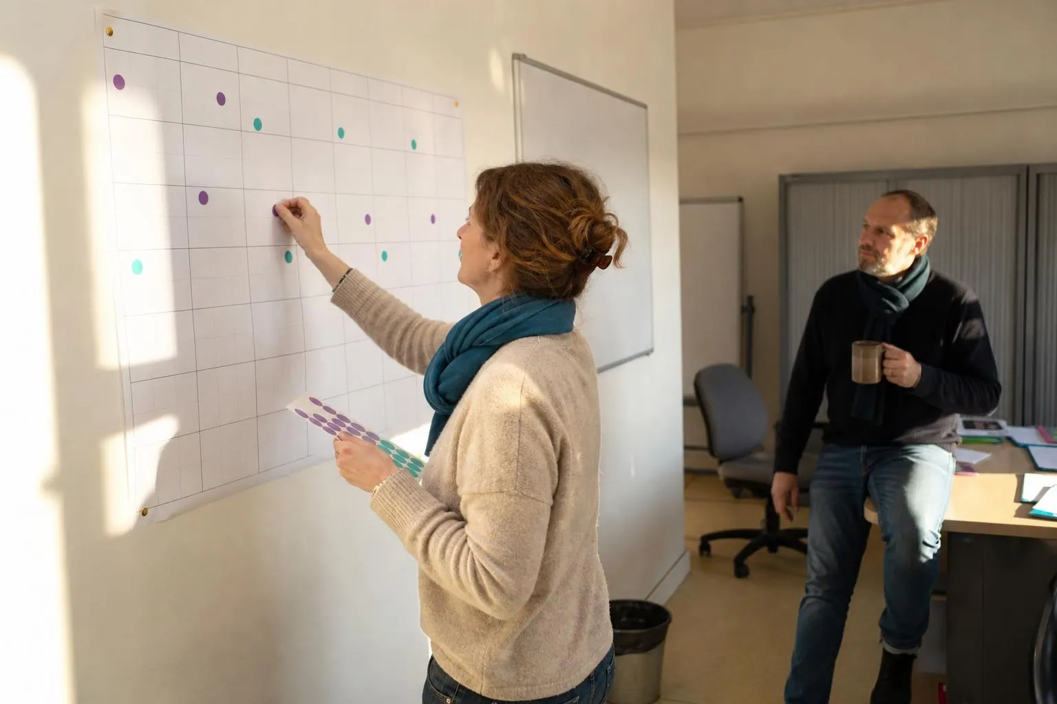 Une responsable administrative pose des pastilles de couleur sur un grand planning annuel papier affiché au mur d’un bureau associatif