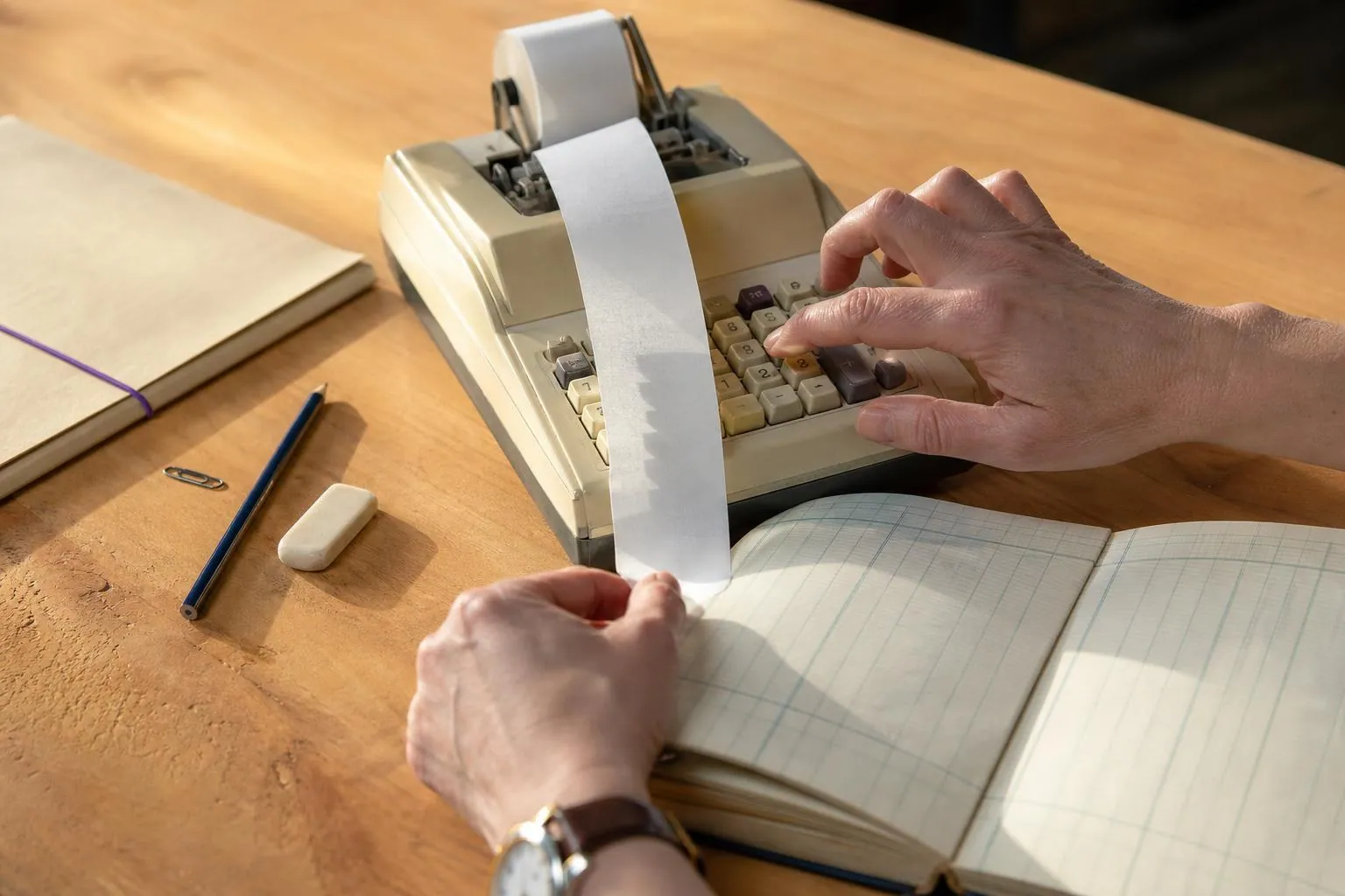 Gros plan sur les mains d’une gestionnaire actionnant une calculatrice à rouleau dont la bande de papier se déroule sur un cahier à colonnes